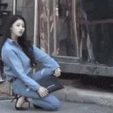a woman in a denim shirt is kneeling down on the sidewalk holding a black purse that says jil sander on it