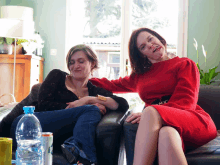 two women are sitting on a couch with one wearing a red dress