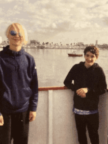 two people standing next to each other in front of a body of water with a boat in the distance