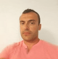 a man in a pink shirt is standing in front of a white wall and looking at the camera .