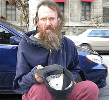 a man with a beard is kneeling in front of a car holding a hat