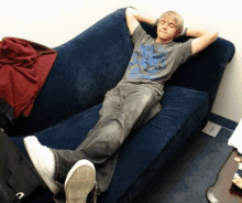 a young man is laying on a blue couch with his legs crossed and his hands behind his head .