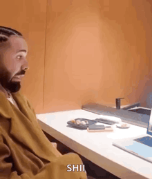 a man with a beard is sitting at a desk with a laptop and a plate of food on it .