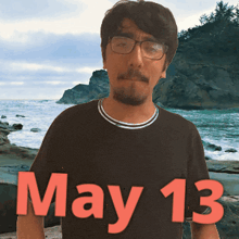 a man wearing glasses stands in front of the ocean with the date may 13 on his shirt