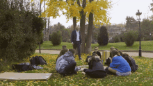 a man in a suit stands in front of a group of people sitting in a circle on the grass