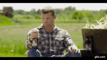 a man in a plaid shirt is sitting on a truck in a field holding a glass of beer .