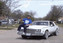 a man in a blue shirt is sitting on the hood of a white car with a license plate that says k78-6030