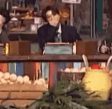 a man in a suit and tie is sitting at a counter in front of a display of fruits and vegetables .