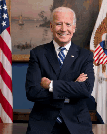 a man in a suit and tie stands with his arms crossed in front of american flags