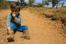 a young boy sitting on a dirt road with a striped shirt and a pair of boots