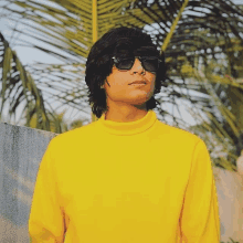 a man wearing sunglasses and a yellow sweater stands in front of a palm tree