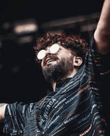 a man with a beard wearing sunglasses and a balenciaga shirt is standing on a stage with his arms in the air .