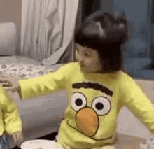 a little girl wearing a yellow sesame street shirt is sitting on the floor .