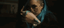 a woman with blue hair and glasses is covering her mouth with her hand in a dark room .