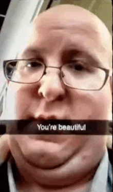 a bald man wearing glasses is making a snapchat with a caption that says `` you 're beautiful '' .