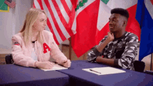 a man and a woman are sitting at a table with flags in the background . the woman has the letter a on her jacket .