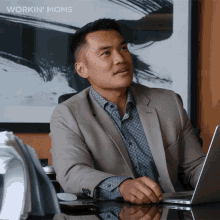 a man in a suit sits at a desk with a laptop in front of him and the words workin ' moms above him