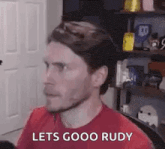 a man in a red shirt is sitting in front of a microphone in a room and says `` lets goo rudy '' .