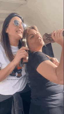 two women are standing next to each other drinking from bottles .