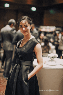 a woman in a black dress stands in front of a table with the website www.niceprintphoto.com on the bottom right