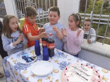 a group of children are gathered around a table with a birthday cake and coca cola bottles