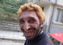 a man with blonde hair and a beard is smiling in front of a building ..