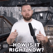 a man with a beard is standing in front of a microphone with the words how is it right now below him