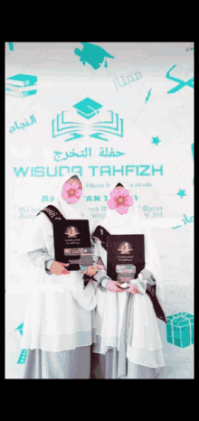 two girls holding their diplomas in front of a sign that says wisuda tahfizh