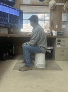 a man sits on a white bucket in front of a computer monitor that shows a graph of a few numbers