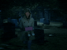 a woman in a trench coat is standing in a cemetery holding a pink box
