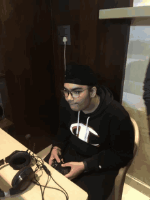 a man wearing a black champion sweatshirt sits at a desk playing a video game