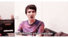 a man in a purple shirt is sitting on a couch talking about destroying a cupcake .