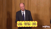 a man in a suit and tie stands at a podium with a sign that says stay protect save the nhs lives