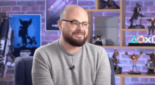 a man with glasses and a beard is sitting in front of a shelf with toys on it and smiling .