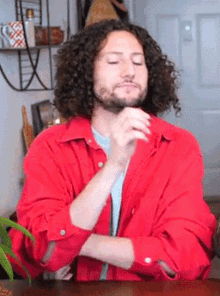 a man with curly hair and a beard wearing a red shirt is sitting at a table with his eyes closed