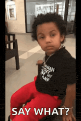 a young boy wearing a black shirt and red pants is sitting in a chair and says " say what " .