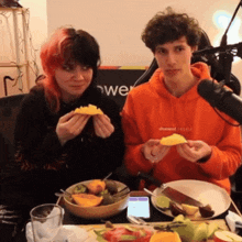 a man and a woman are sitting at a table eating fruit and one of them is wearing an orange hoodie with the word channel on it