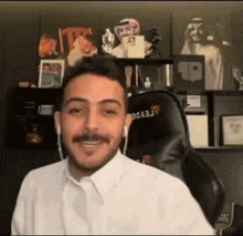 a man with a mustache is smiling while sitting in a chair in front of a shelf with pictures on it .