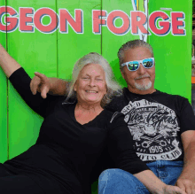 a man and a woman are posing in front of a green sign that says geon forge