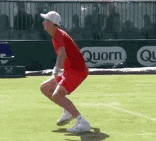 a man in a red shirt and white hat is jumping on a tennis court with a quorn ad in the background