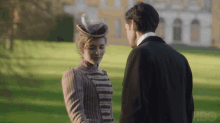 a man and a woman are looking at each other in a park . the woman is wearing a feathered hat .