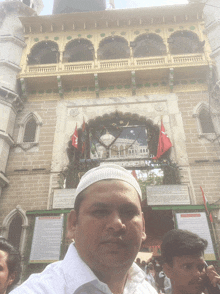 a man wearing a white hat stands in front of a building that says ' masjid '