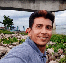 a man in a blue shirt smiles in front of a bridge over a body of water