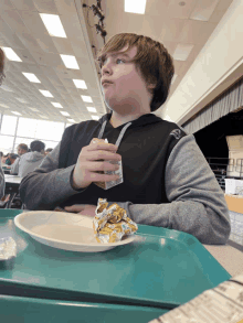 a boy is sitting at a table in a cafeteria holding a carton of milk