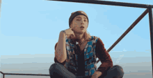 a young man wearing a beanie and a plaid shirt is sitting on a railing looking at the camera .