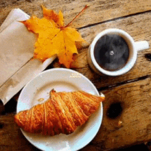 a croissant on a plate next to a cup of coffee and a maple leaf on a wooden table .