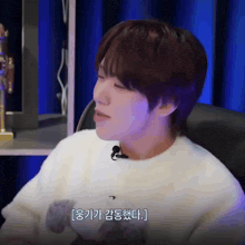 a man wearing a white sweater with a teddy bear on it is talking into a microphone while sitting in a chair
