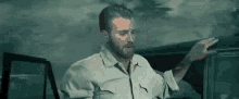 a man with a beard is standing in front of a car and pointing at it .