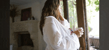 a woman in a white robe looks out a window holding a cup of coffee