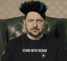 a man wearing a black shirt that says stand with redan sits in a chair